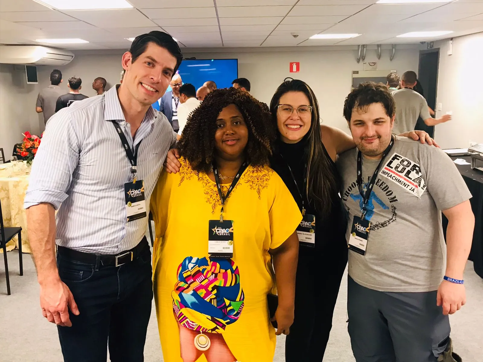 Camila Georg, de vestido amarelo, abraçada a três participantes do CPAC Brasil, todos com crachá do evento.