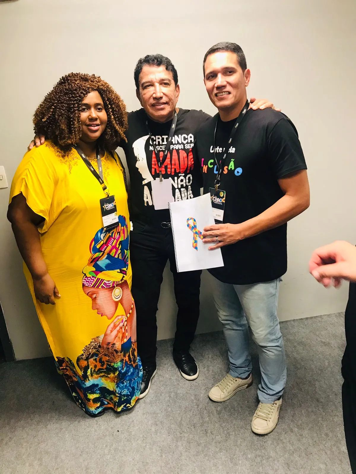Camila Georg, de vestido amarelo, ao lado do senador Magno Malta e de um homem que segura um caderno com a fita do autismo, todos com crachá do CPAC Brasil.