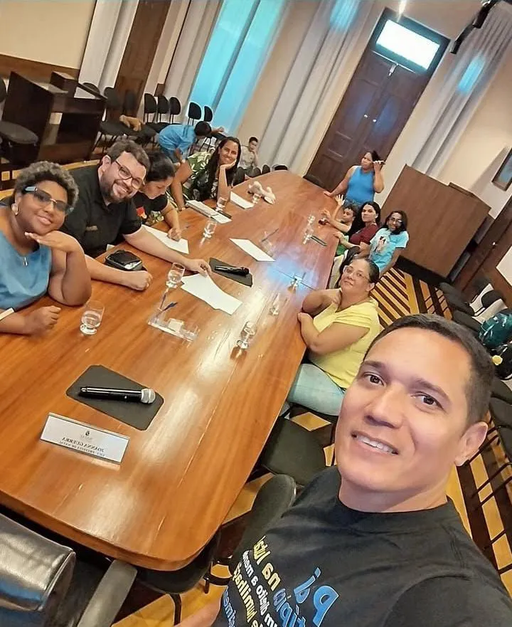 Selfie de um pai em uma mesa de reunião comprida: mães, pais e crianças sentados ao redor, com copos d'água, papéis e microfones.